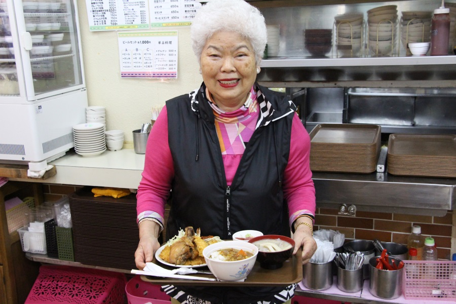お盆にのせた料理を手に、ニッコリ微笑む店のアンマー